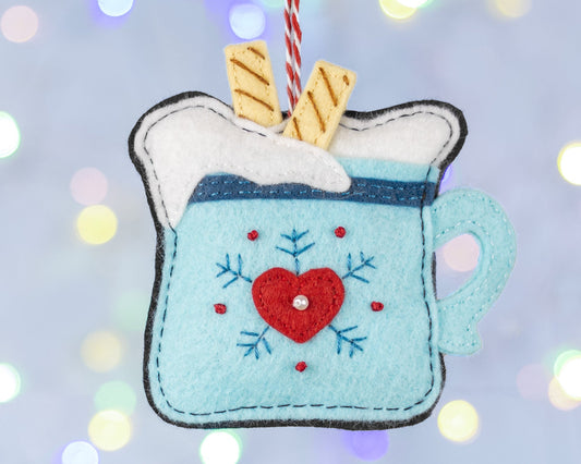 A hot chocolate felt ornament. The light blue mug has a red heart and snowflake decoration, the top has a layer of cream with 2 wafer biscuits. The ornament is hanging with blurred fairy lights in the background.