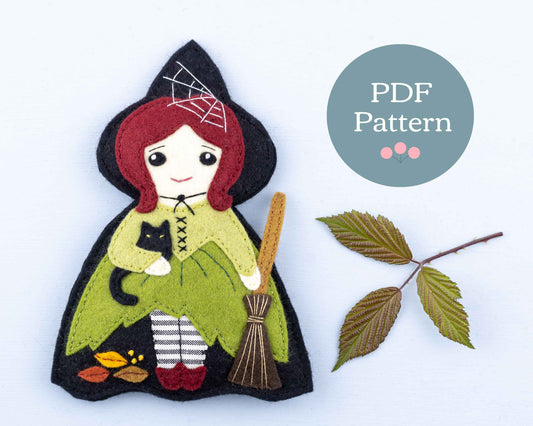 A felt witch ornament with red hair and a green dress. She has a black pointed hat and cloak, carries a broom and has a black cat tucked under her arm.
