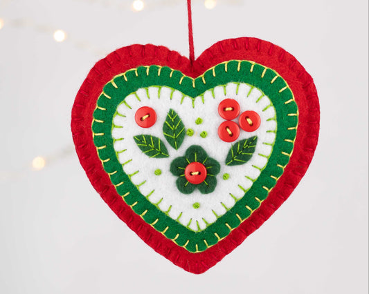 A felt heart ornament with layers of red and green and a white centre with a flower, holly leaves and button berries.