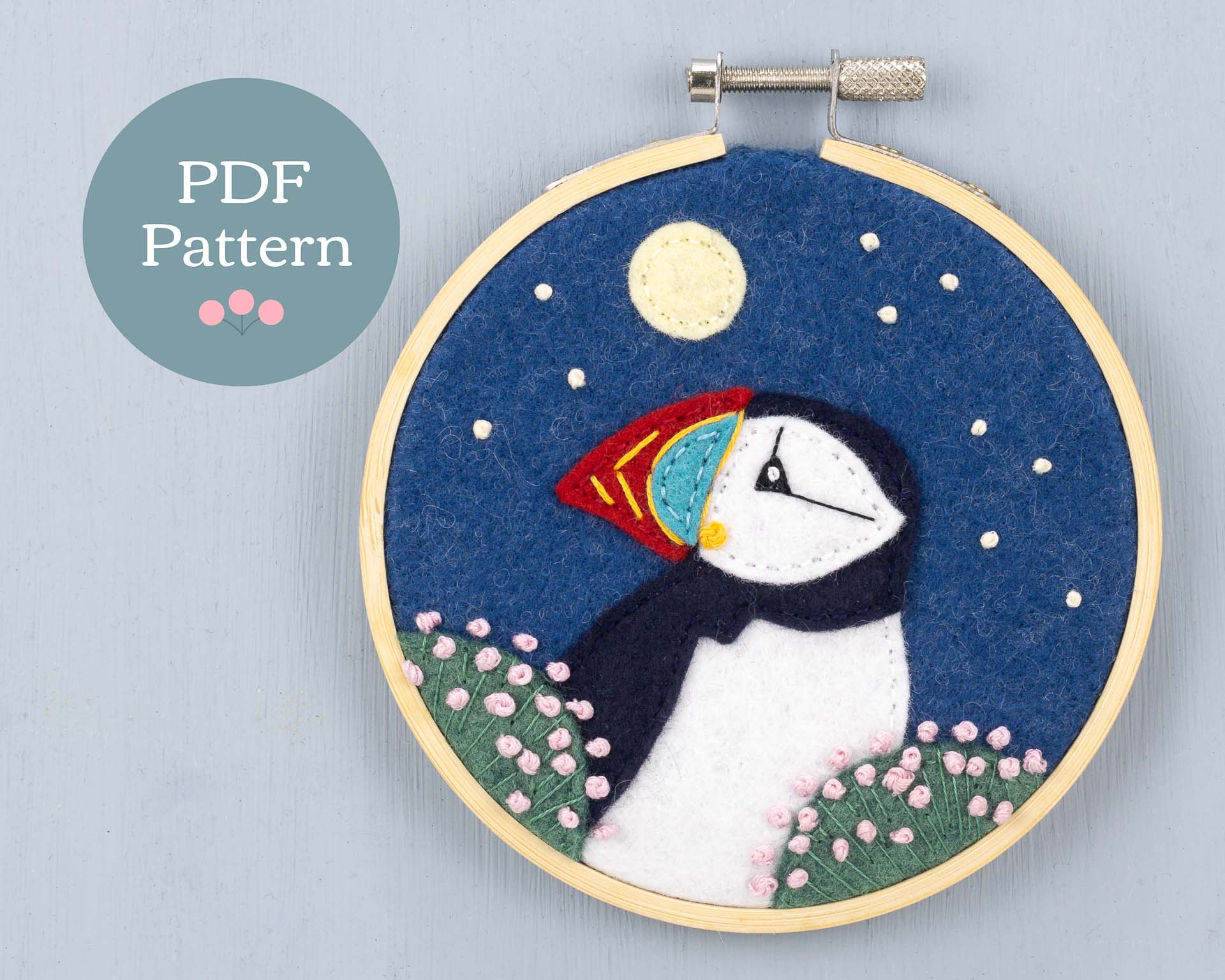 Felt applique and embroidery showing head and shoulders of a puffin among clumps of sea pink flowers. The background is a starry sky with a full moon. Framed in a wooden embroidery hoop.