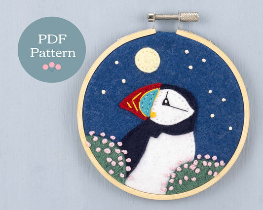 Felt applique and embroidery showing head and shoulders of a puffin among clumps of sea pink flowers. The background is a starry sky with a full moon. Framed in a wooden embroidery hoop.