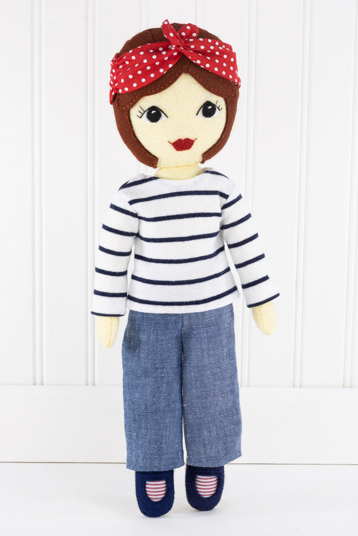 Tilly standing against a white background. She wears a white top with navy stripes, denim pants, navy shoes and a red and white polka dot bandana.