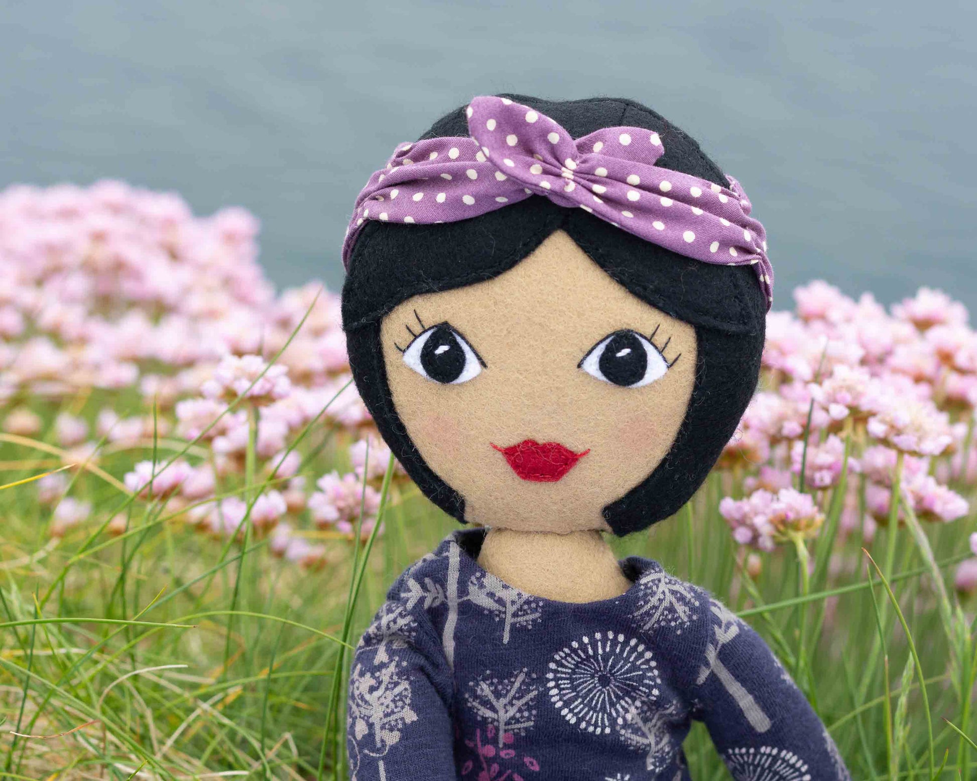 A Tilly doll with black hair and medium skin tone wearing a purple and white polka dot bandana and a floral patterned top, with sea pink flowers and the sea in the background.