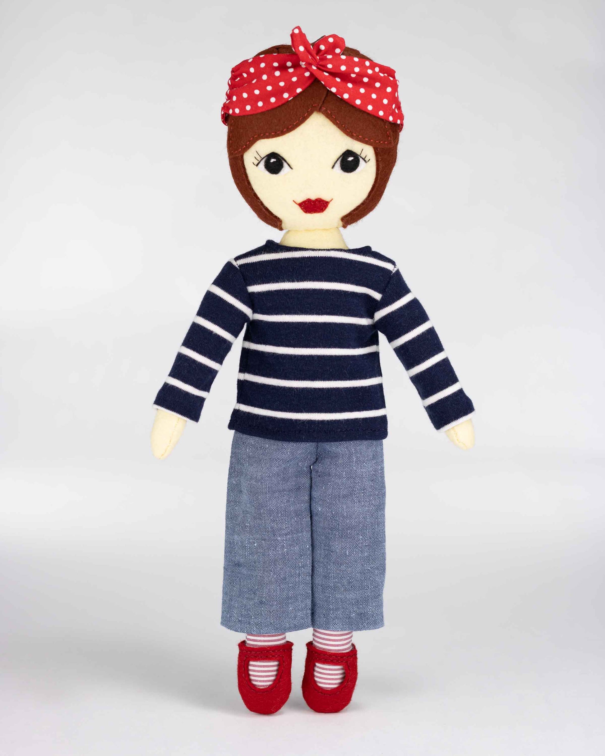 Tilly standing against a white background. She wears a navy top with white stripes, denim pants, red shoes and a red and white polka dot bandana.